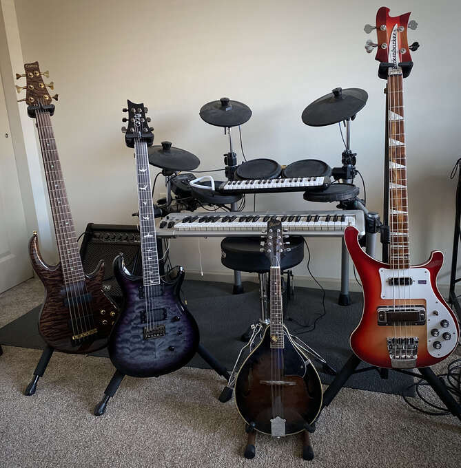 A six-string bass, a four-string bass, a guitar, and a mandolin on stands in front of an electronic drum kit. A MIDI keyboard and a melodica rest on the drum throne.