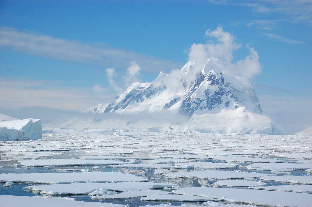 A snow-covered mountain with fog around it in the background and sheets of sea ice packed densely in the foreground.