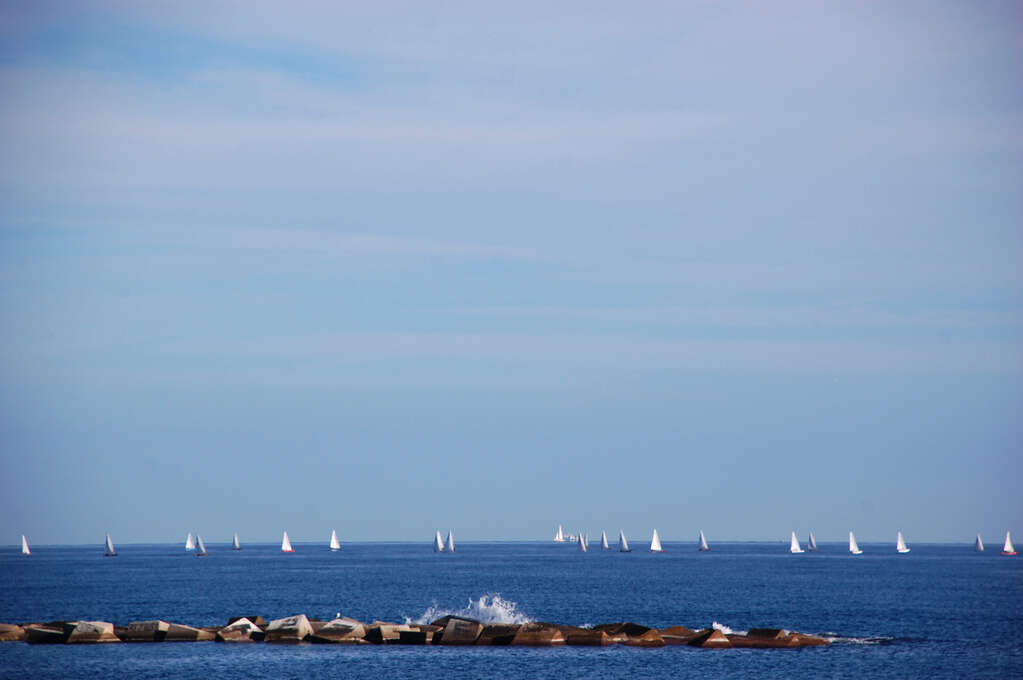 A sea with many sailboats and a rocky pier with a crashing wave.