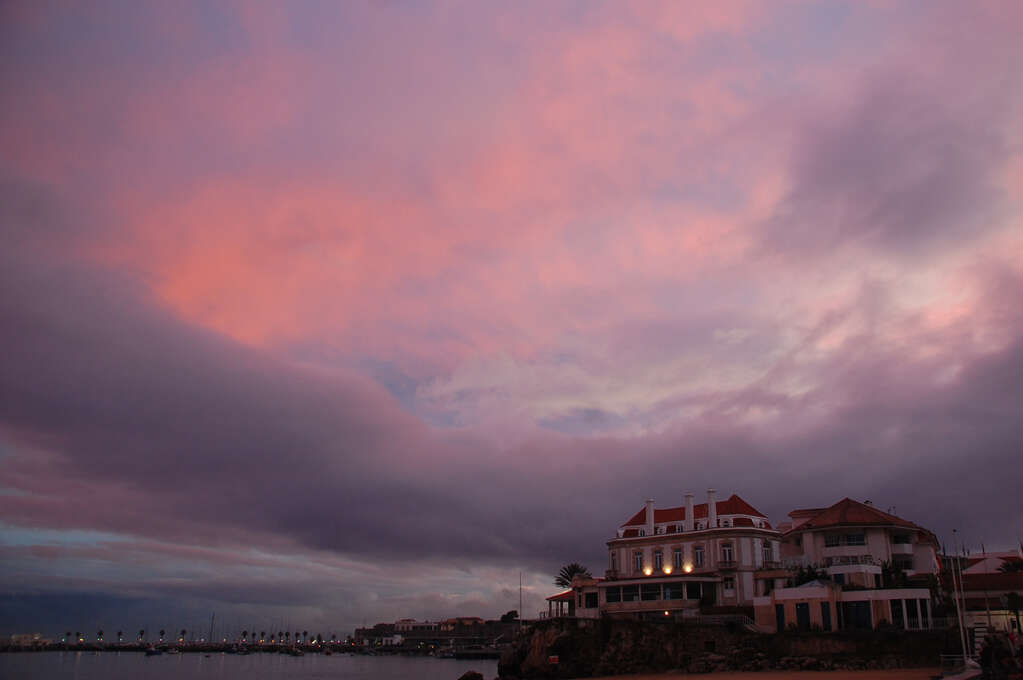 A cloudy sky colored by the sunrise and a large house with lights in the front.