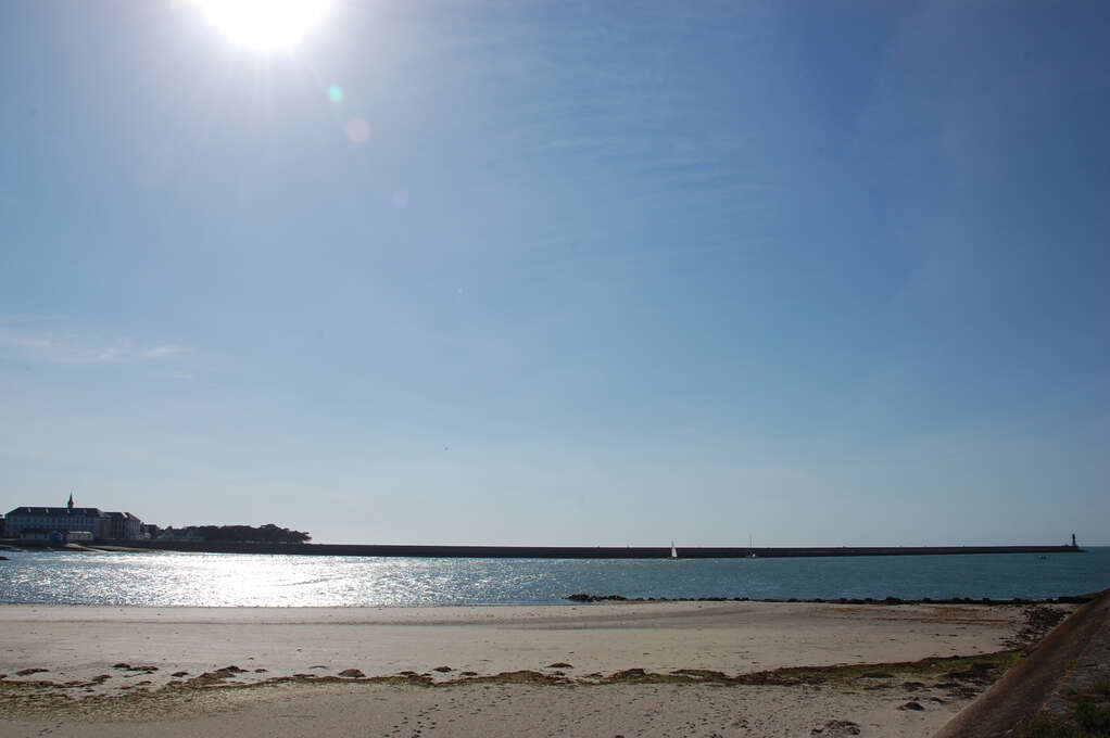 A sunny beach with a long pier in the background leading to a small lighthouse.