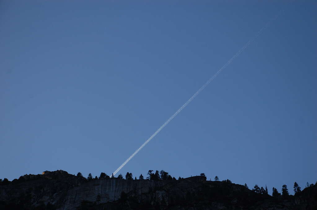 A jet trail angled down towards a cliff and disappearing behind it.